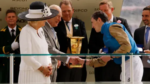 Reuters James Doyle stands to the right, he is slightly bowing as he shakes King Charles' hand, who is standing to the left and Queen Camilla is standing next to King Charles. Doyle is still in his racing outfit, a blue bomber style jacket with tan coloured sleeves and white bottoms. King Charles wears a light grey suit with a white shirt and pale blue tie. Queen Camilla wears a smart white long sleeve dress with a pleated skirt, with a brooch and black and white wide brimmed hat which has a cluster of flowers on the headband. King Charles is smiling as he shakes Doyle's hand and passes him a golden trophy. There is a row of mean in suits in the background all smiling watching the interaction.