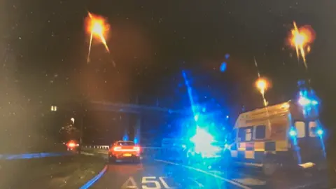Ambulances and a police car are pulled up on a lane of a dual carriageway. It is dark, and their blue lights are reflecting off the camera