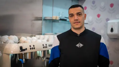 A man in his 20s with short black hair looks at the camera. He is standing in a cafe, next to a coffee machine with mugs stacked on top of it. He is wearing a black jumper with blue and white patches on the sleeves. 