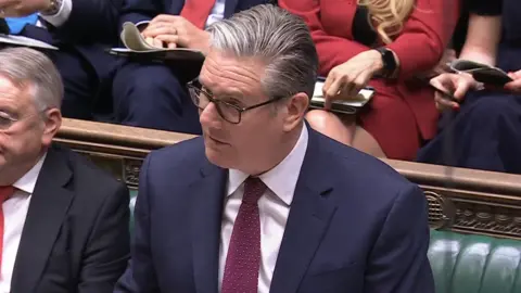 House of Commons Sir Keir Starmer is standing in the House of Commons. He is in front of a green bench and has short greying hair, is wearing black framed glasses, navy jacket, pink shirt and purple patterned tie.