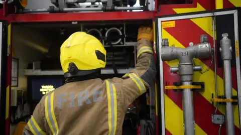The backs of three firefighters wearing breathing equipment and helmets. A fire station and other firefighters are out of focus in the background.
