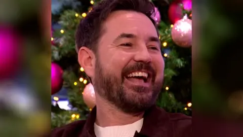 Martin Compston laughs in front of a decorated Christmas tree