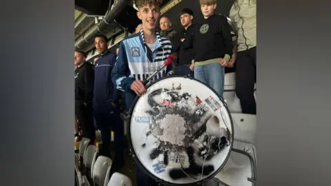 Corey Matthews Cory stands next to his damaged drum. The skin is ripped, with the remnants showing the stickers fans have affixed to it over the years. Cory is wearing a Coventry City hooded top with 'Hummel' on the chest.