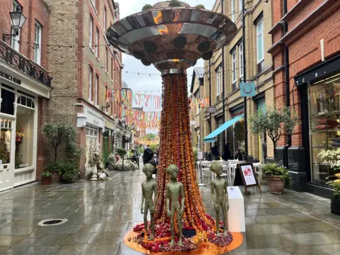 A tall, flower-covered column topped with a metallic UFO disc stands in a narrow street, surrounded by three small green alien figures and colourful bunting strung between brick buildings.