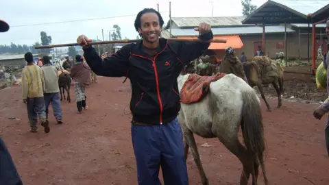 Jerry Rothwell Image shows Dan with a black zip up hoodie with red detailing. He is wearing blue trousers. He has a stick across his shoulders. His hair is pushed back away from his face. He is in Ethiopia. Behind him is a horse with a small rug over it's back. It's got white fur. The floor is a red in colour. 