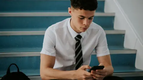 an adolescent boy in a short-sleeved shirt and a tie sitting on stairs, looking at a phone