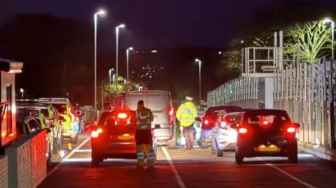 IOM CONSTABULARY Cars line up in pit lane at the grandstand at night with the rear lights illuminated. There are officers in hi-visibility jackets walking between them