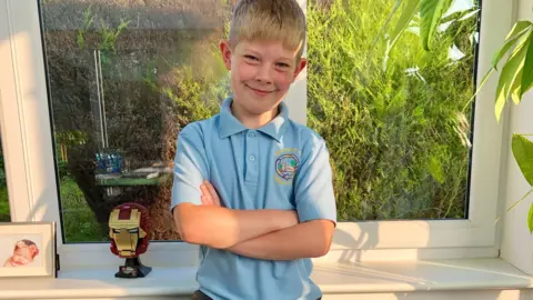 An eight-year-old boy stands with his arms folded in his school uniform, at home by a window