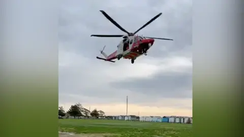 Supplied A red and white helicopter taking off from a field with beach huts in the background