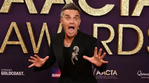 Robbie Williams is making a surprised face at an awards ceremony. He is wearing a black suit jacket and black t-shirt and has dark hair spiked. 
