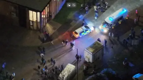A drone shot of a city street at night time. A police car and a police van are visible and a number of people are milling about. Two white vans are also visible, and light is spilling from the windows of one building. 