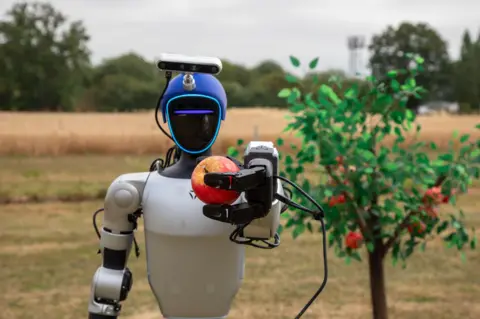 A humanoid robot holds a red apple it has just picked from a tree.