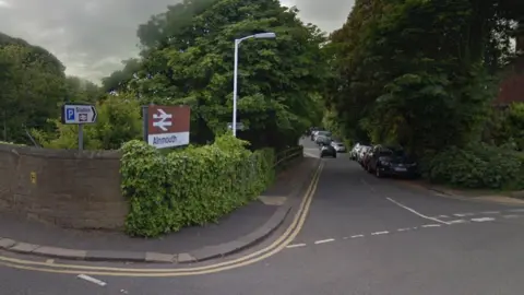 A street view with a sign pointing to Alnmouth train station. Cars and trees line the road.