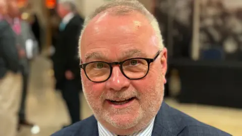 Shariqua Ahmed/BBC Wayne Fitzgerald smiling for the camera. He has short white hair and is wearing a blue-and-white striped shirt and a blue blazer.