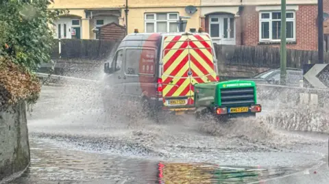 BBC WEATHER WATCHERS/STORMCHASERLIAM A van drives through floodwater with houses visible in the background