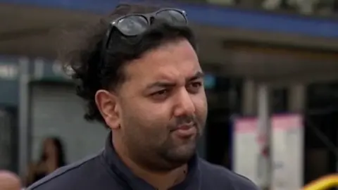 A man wearing a navy polo shirt. He has short curly hair and is wearing sunglasses on the top of his head.