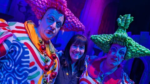 The owner and manager of The Academy Theatre in Birdwell stands between two panto dames who are wearing brightly coloured costumes and exaggerated make-up. The dame on the left is wearing a bright pink pointy plastic wig. The dame on the right is wearing green pointy plastic hair.
