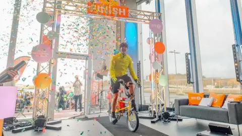 BBC A smiling Paddy McGuinness in a bright yellow top riding his yellow bike through a large metal frame bearing the word "finish", as confetti falls around him