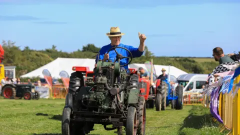 CALLUM STALEY/CSJ PHOTOGRAPHY A man driving an old fashioned tractor wearing a trilby hat and giving the watching a thumbs up. He is leading a parade of other tractors in the grassed show ring.