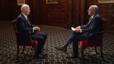 Joe Biden wears a dark blue suit and sits in a red chair in a room with a patterned carpet and wood-paneled walls. He faces the BBC's Nick Robinson, who also wears a dark suit and sits in a chair.