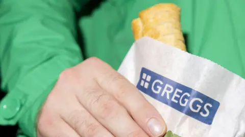 Getty Images A person in a bright green jacket holds a Greggs paper bag with a sausage roll peeking out from the top, the blue-and-white Greggs logo clearly visible on the front.
