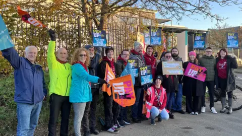 A group of teachers standing outside Barton Peveril college in Eastleigh wearing hats and gloves. They're holding banners saying Pay Up.