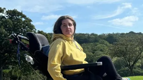 A woman using a wheelchair is wearing a yellow jumper and looking straight to camera. There is greenery in the background.