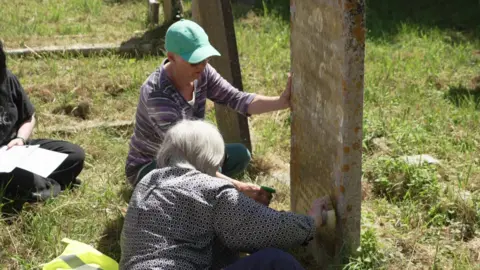 Two women with white hair are scrubbing a headstone to make it readable. The grass surrounding the grave is dry.