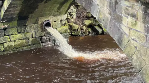 A combined sewer overflow pipe discharging fast flowing water into the River Calder at Hebden Bridge. The water looks comparatively clean and is likely to be a mixture of rainwater and wastewater.