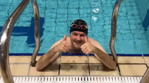 BBC Michael Coombes, a man wearing a swimming cap and leaning on the edge of an indoor pool, gives a thumbs up.