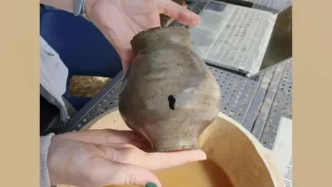 University of Leicester A woman's hands holding up a Roman pot with a hole in its side over a bowl of muddy water