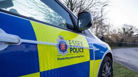 Essex Police car parked by the scene of a crash. It has logo saying Essex Police and. The car is painted in neon yellow and blue checks.