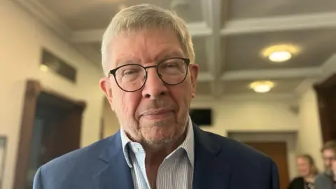 Ross is wearing a blue blazer and white and blue stripped shirt.