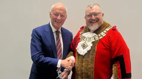 Brentwood Borough Council Barry Hearn, wearing a navy suit jacket, white shirt and red and blue striped tie, shaking the hand of Jay Laplain, who is wearing a red ceremonial mayor's outfit.