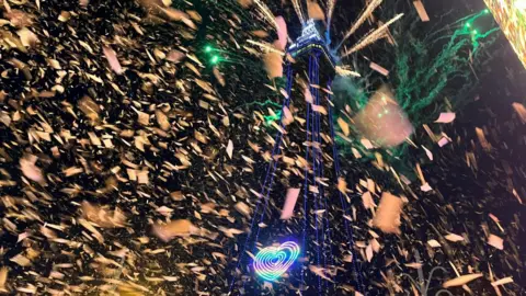 BBC / Zoe Toase Blackpool Tower is lit up with blue lights and a rainbow-coloured heart and ticker tape. Green fireworks can be seen behind it.
