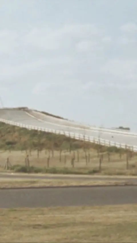 A large, grey ski slope located right on top of a hill and by the side of the motorway