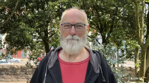 Nigel Jones smiles at the camera. He has glasses and a white beard. Is wearing a red tshirt and black hoody. Trees visible in the background.