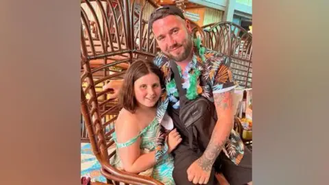 Conaire Quinn Conaire Quinn with his daughter Autumn. Conaire is sitting on her lap at a seating area. Autumn has short brown hair and is wearing a blue print dress. Conaire is wearing a hawaiian t-shirt. He has short brown hair and a beard and is wearing a baseball cap (on backwards) and a floral garland around his neck.