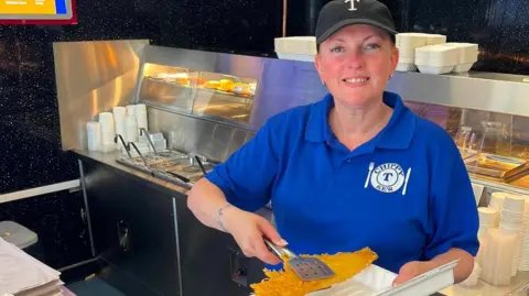 Tracy Stock with hair tied back wearing black baseball cap and a blue t-shirt stands with a white Chippy-T logo stands in front of the frying range in a fish and chip shop holding a battered fish over a take out container with stainless steel tongs. She is smiling.