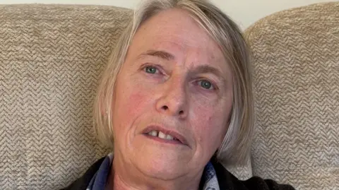 BBC A woman with grey hair sitting on a light sofa