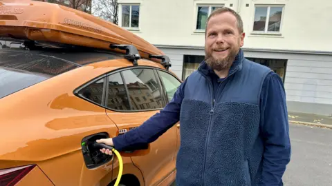 Pengendara BBC Norwegia Ståle Fyen tersenyum sambil memasang kabel pengisi daya ke mobil listriknya