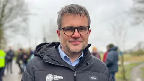Tom Jackson/BBC Craig Bennett has short grey hair and beard and is wearing black framed glasses. He is wearing a blue shirt with a black coat which has a logo of a badger and says 'The Wildlife Trusts'. People are standing behind him. 