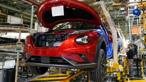 Reuters A car up in the air on a conveyor belt. A man is working on the car behind