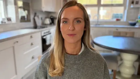 BBC Natasha Murray has balayaged hair and a grey jumper. She sits in her kitchen looking into the camera in the centre of the frame. 