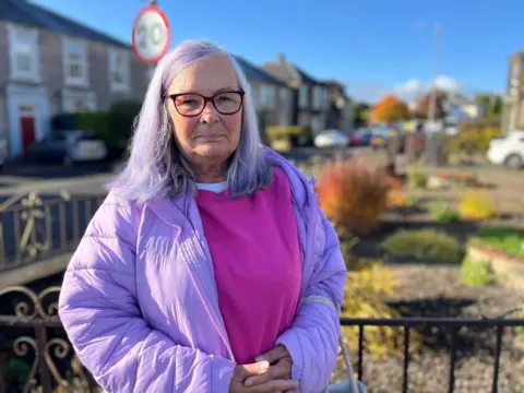 A woman wearing a purple top and jacket standing in a residential street looking at the camera