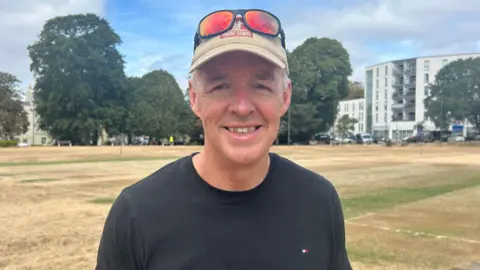 Bill Martin from Plymouth Cricket Club is stood on the playing surface and is smiling looking straight at the camera. He is wearing black t-shirt, beige cap with sunglasses rested on it. The field behind him is parched and dry.