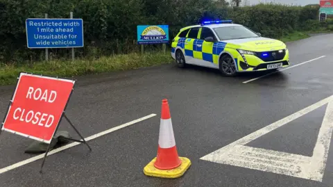 A police car with blue flashing lights parked across a road junction. There is a sign reading 'road closed', a traffic cone and a traffic sign 
saying the road is unsuitable for wide vehicles. 