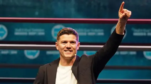 Getty Images Luke Campbell standing in front of a turquoise background with his left hand raised in the air