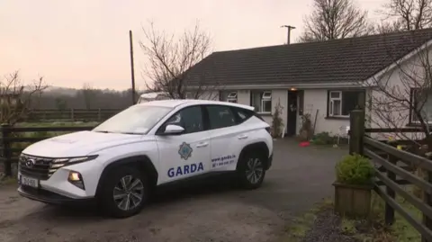 RTÉ News A Garda car sits in front of a white bungalow. A Christmas decoration hangs from the front door. A pair of little trees sit on each side of the front door. 