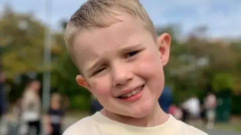 Ozzy, a young boy with blond hair, stands smiling with his head titled to the side. He is wearing a cream coloured t-shirt.